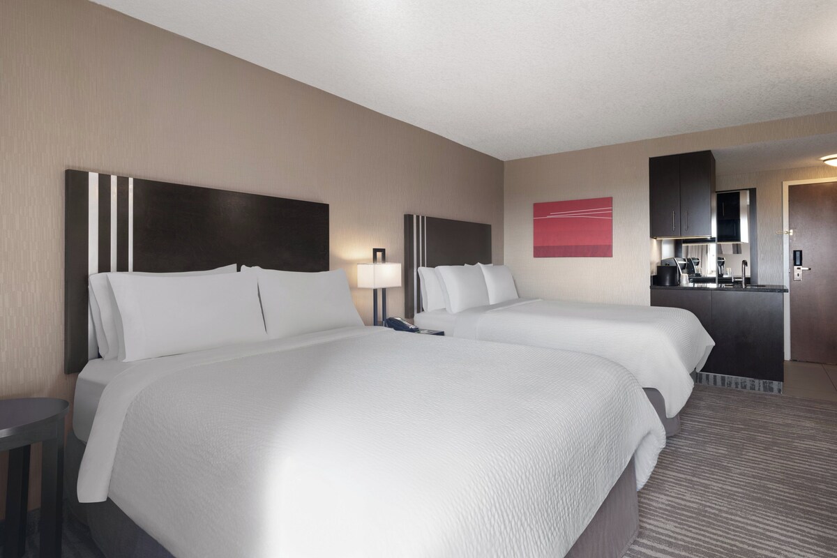 A hotel room with two beds, a desk, a lamp, a mirror, a red artwork, and a kitchenette.