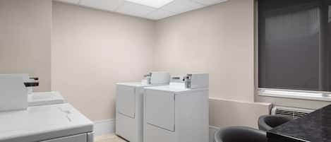 A laundry room with two white washers and dryers, a black chair, and a window with blinds.
