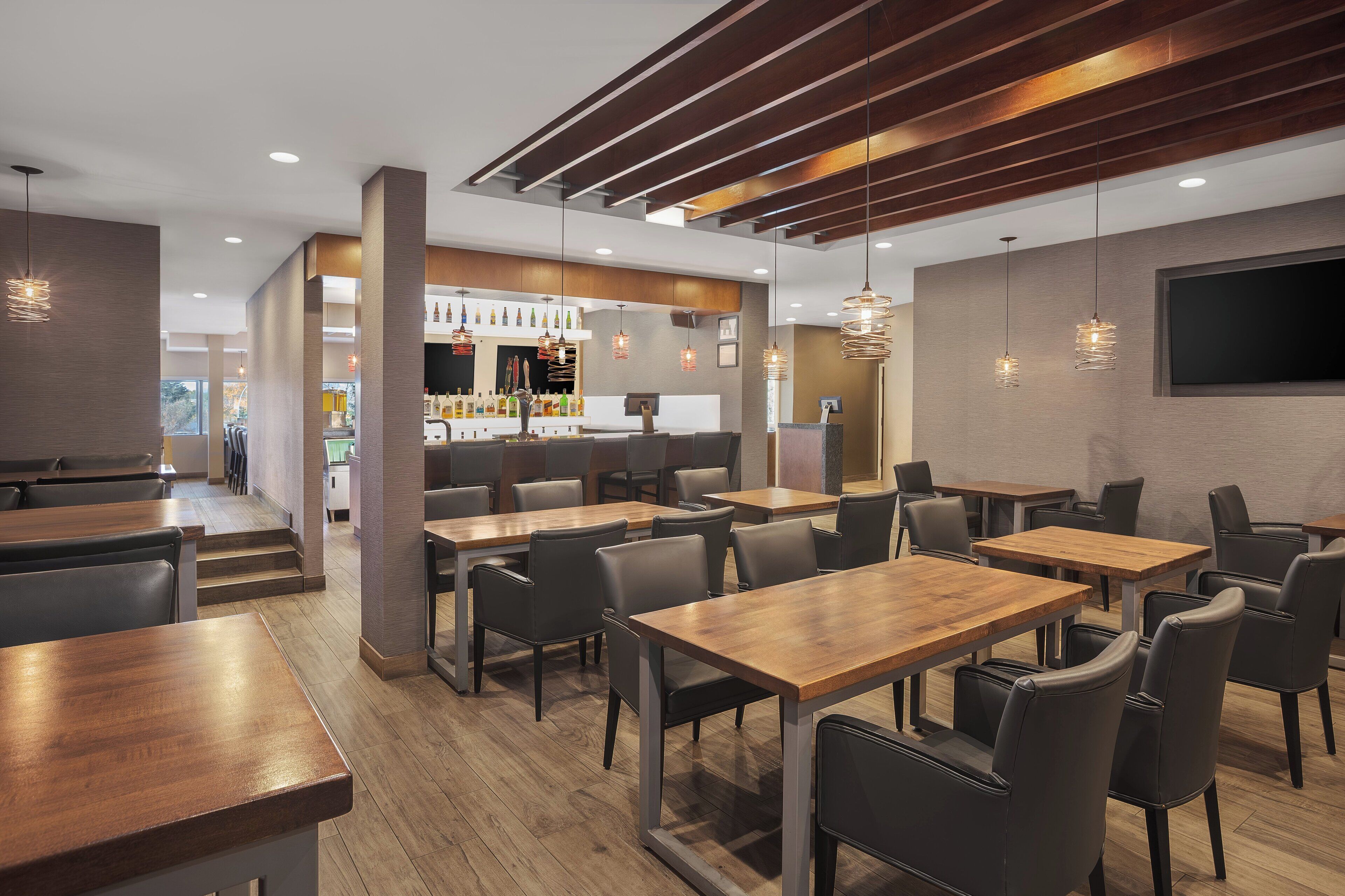 A modern restaurant with wooden tables and chairs, a bar area, and pendant lighting.