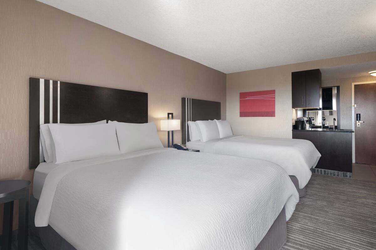 A hotel room with two beds, a desk, a lamp, a mirror, a red artwork, and a kitchenette.