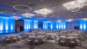 A large banquet hall with round tables and chairs, blue ambient lighting, and modern ceiling fixtures.