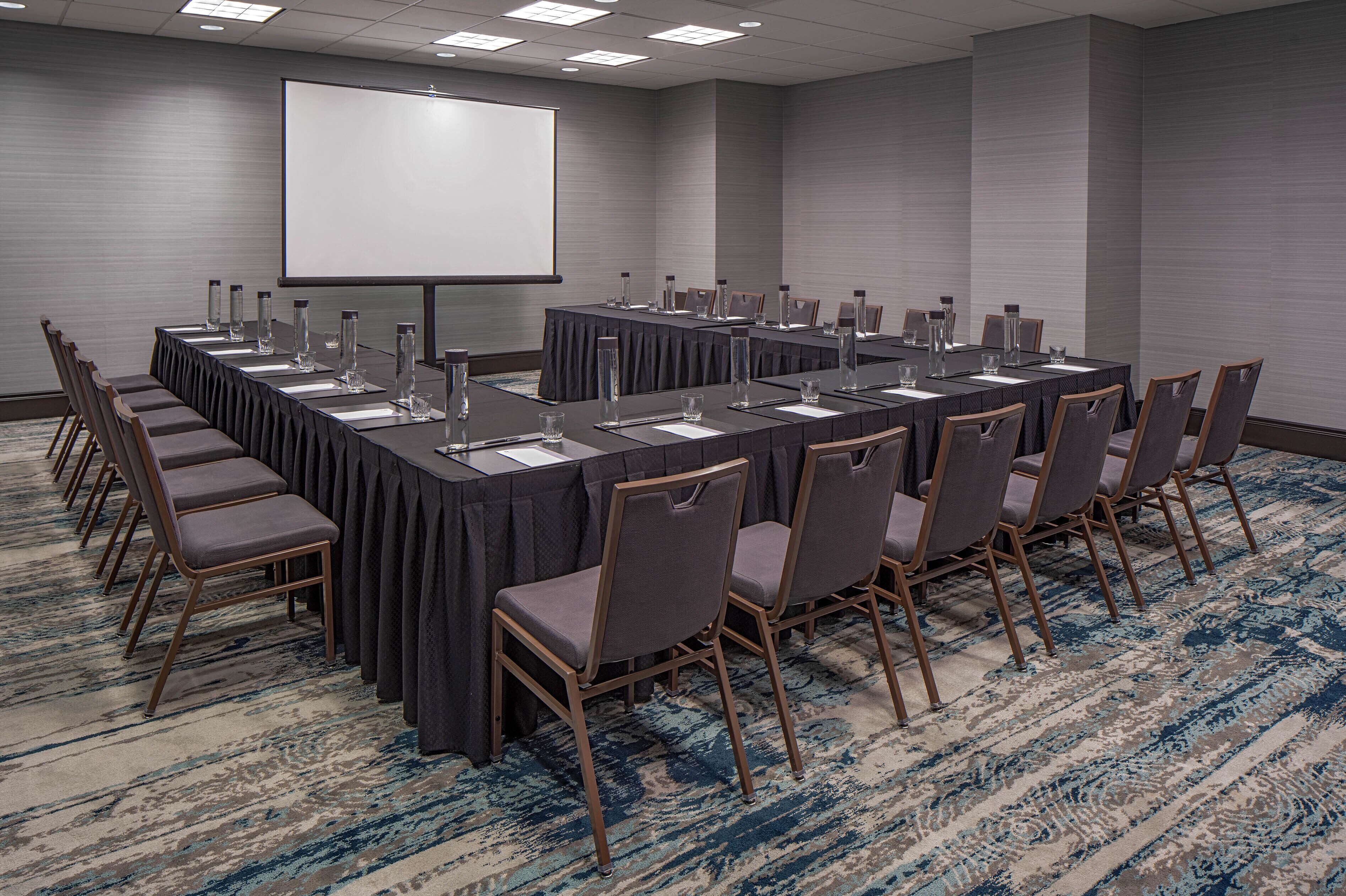 A conference room with a long table, chairs, a projector screen, and a carpeted floor.