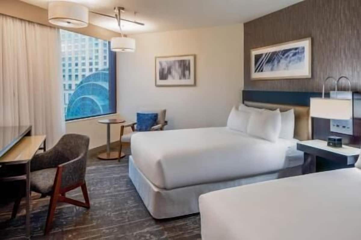 Une chambre d’hôtel avec deux lits, un bureau, une chaise, une petite table et une vue sur un immeuble par la fenêtre.
