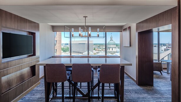 A modern conference room with a large table, four chairs, a flat-screen TV, and a chandelier.