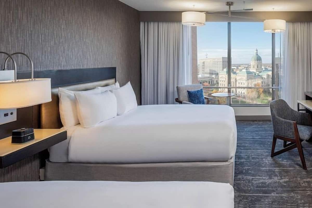 Une chambre d’hôtel avec deux lits, un bureau, une chaise, une lampe et une vue sur le paysage urbain.