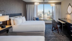 A hotel room with two beds, a desk, a chair, a lamp, and a view of a cityscape.