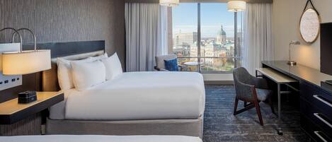 A hotel room with two beds, a desk, a chair, a lamp, and a view of a cityscape.