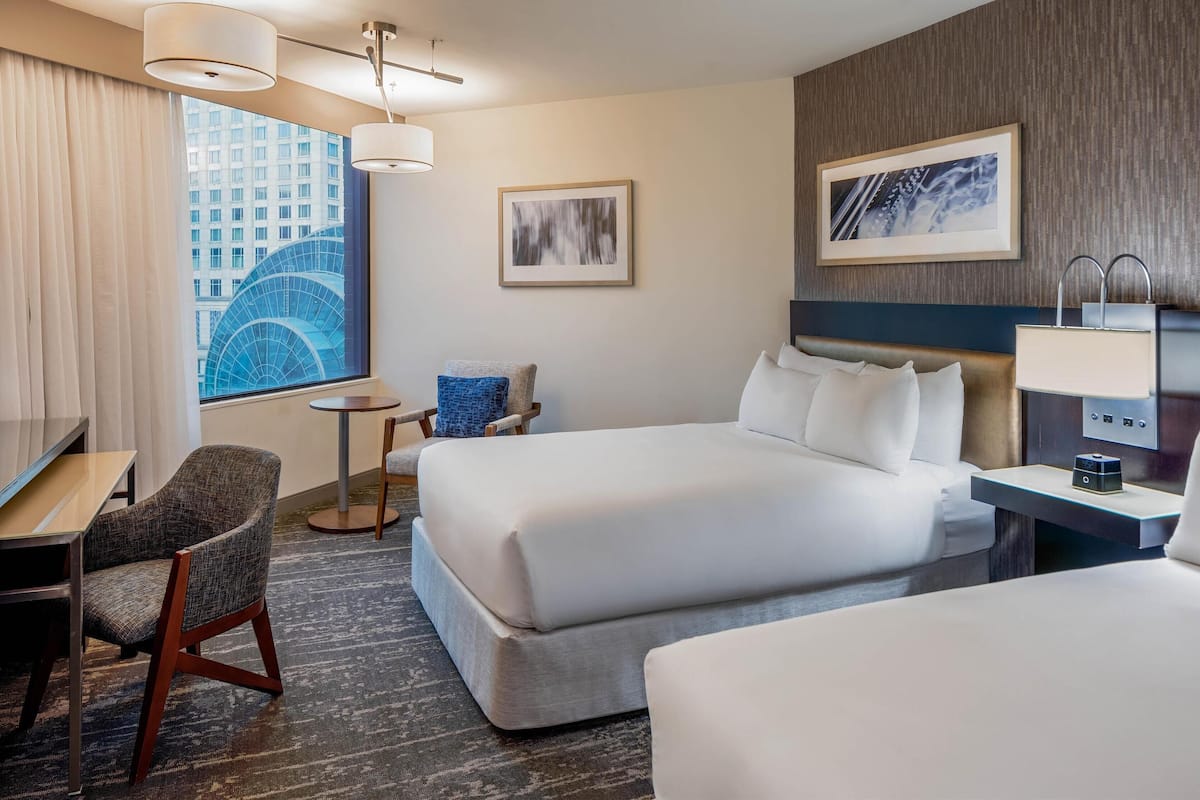 Une chambre d’hôtel avec un grand lit, un bureau, une chaise, une petite table et une vue sur le paysage urbain par la fenêtre.