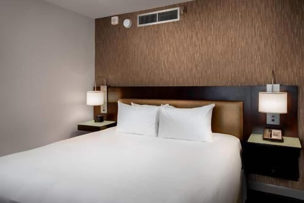 Une chambre d’hôtel avec un grand lit, deux tables de chevet avec des lampes, une tête de lit en bois et une grille d’aération fixée au mur.