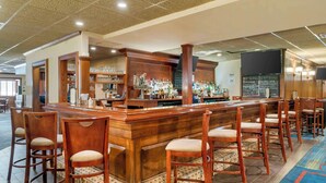 Zona de bar con mostrador de madera, varios taburetes y una variedad de botellas y vasos en estantes.