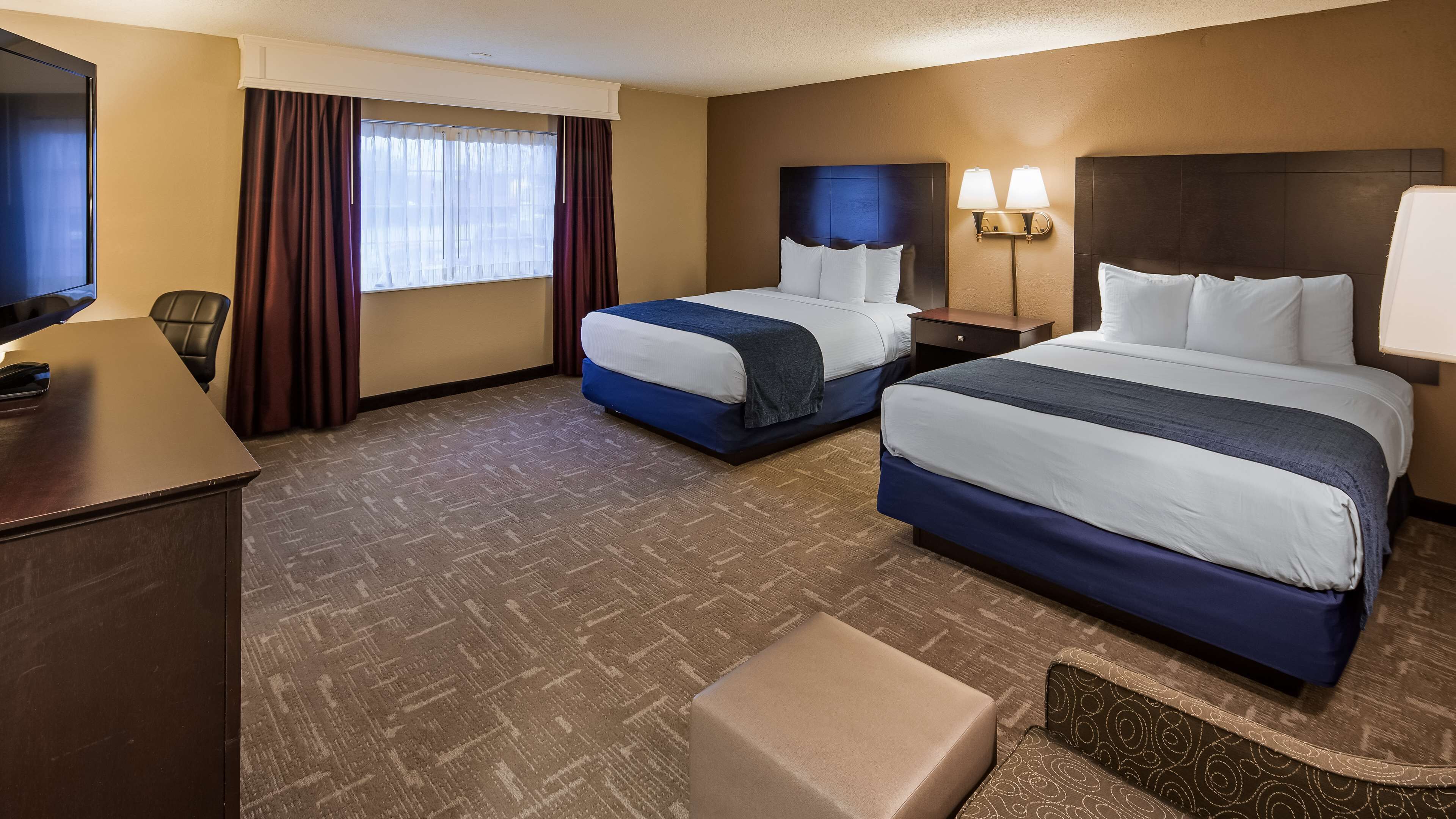 A hotel room with two beds, a desk, a TV, and a window with curtains.