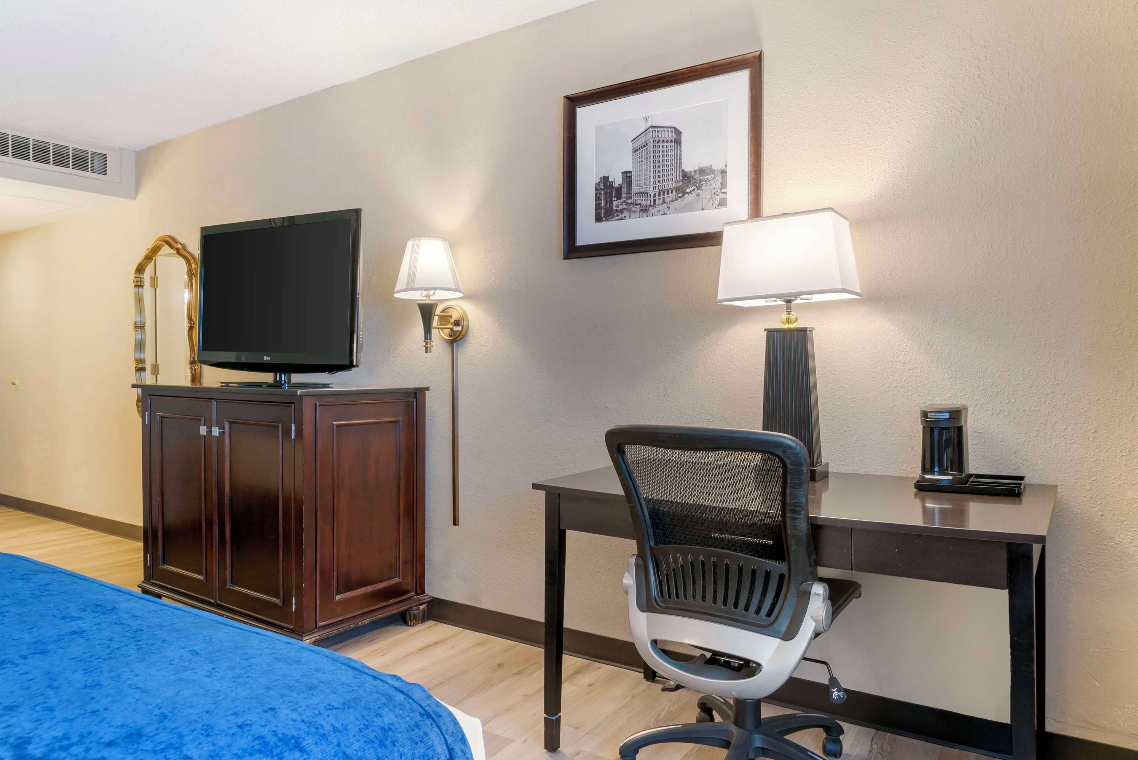 Une chambre d’hôtel avec un lit, un bureau et une chaise, une télévision, un tableau encadré au mur et une lampe.