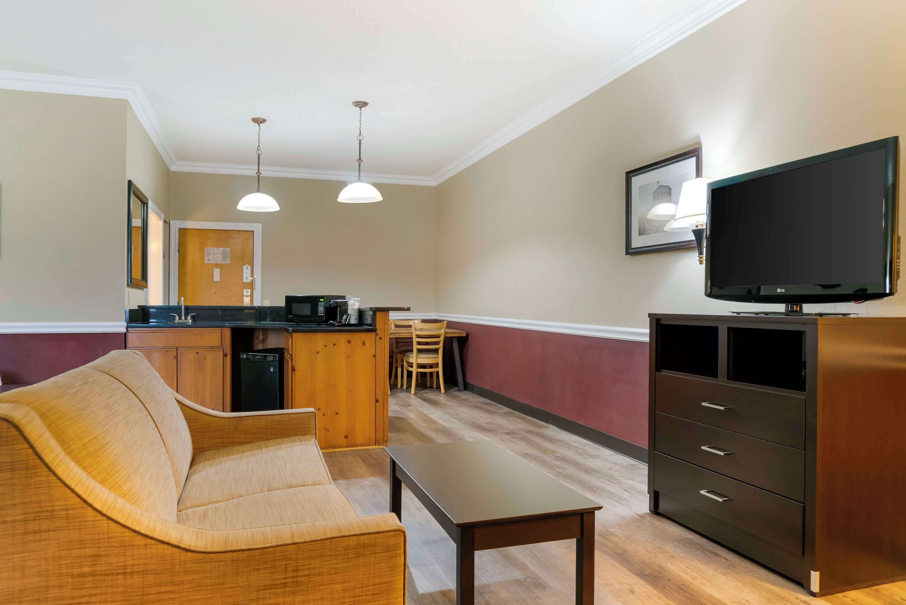 A hotel room with a sofa, a small table, a TV, a kitchen area with a counter and chairs, and a wooden cabinet.