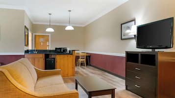 Ein Hotelzimmer mit einer Couch, einem kleinen Tisch, einem Fernseher, einer Küchenzeile mit einer Theke und Stühlen sowie einem Holzschrank.