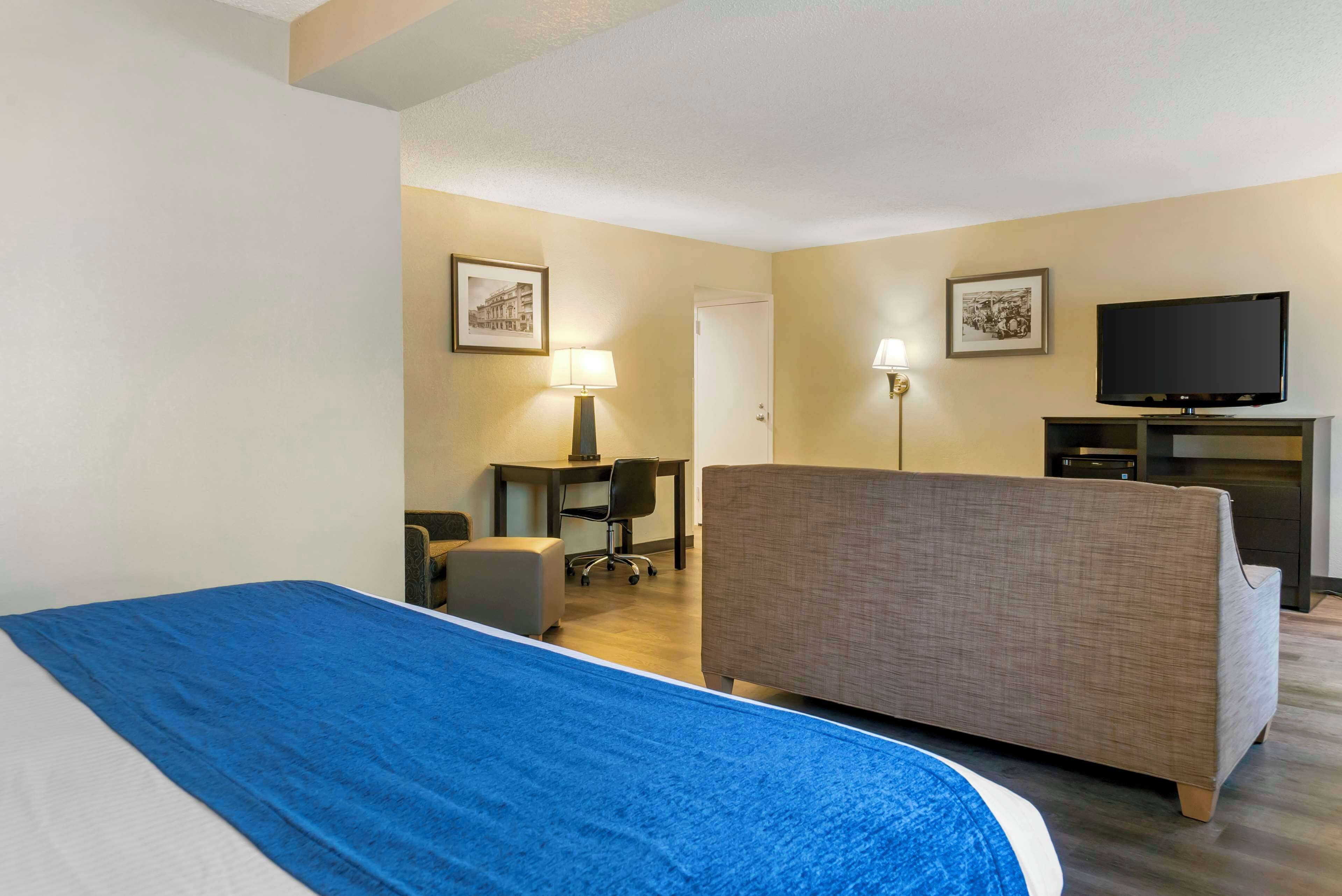 A hotel room with a bed, a sofa, a desk with a chair, a television, and framed pictures on the walls.