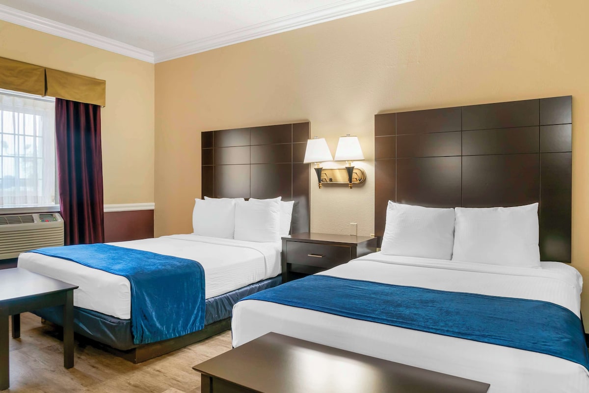 A hotel room with two beds, a nightstand with lamps, a window with curtains, and an air conditioning unit.