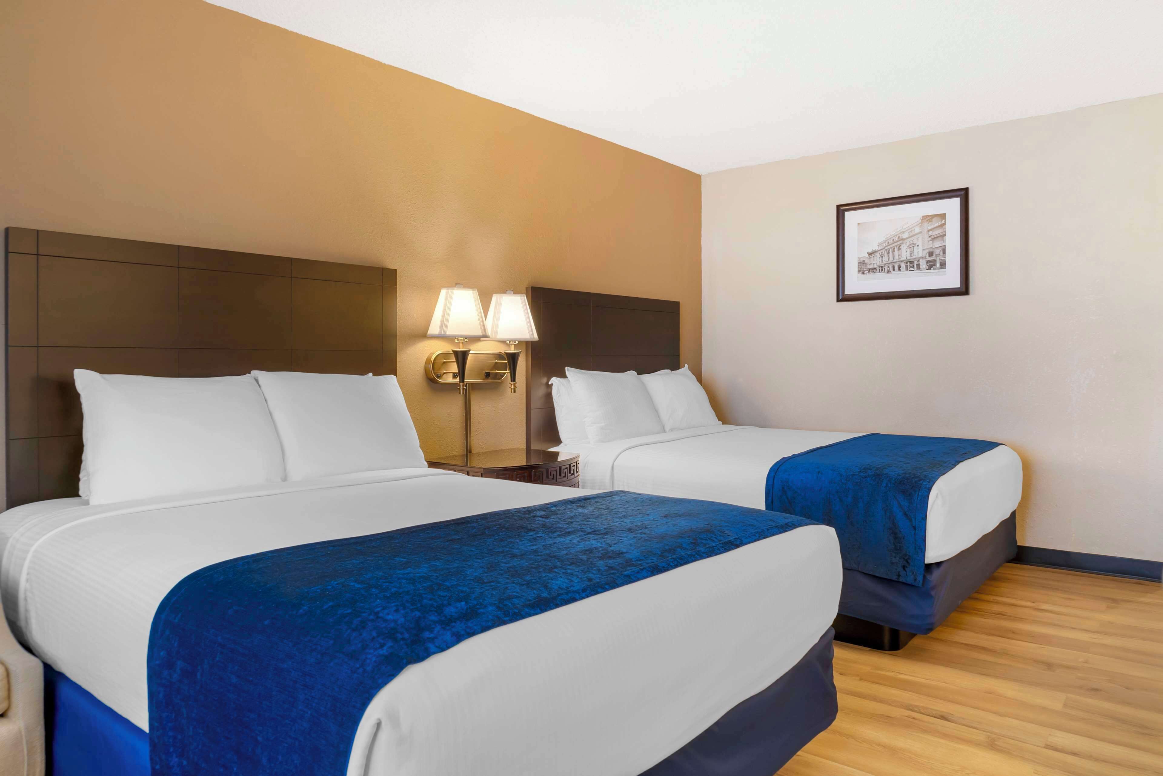 A hotel room with two beds, a headboard, bedside lamps, a framed picture on the wall, and wooden flooring.