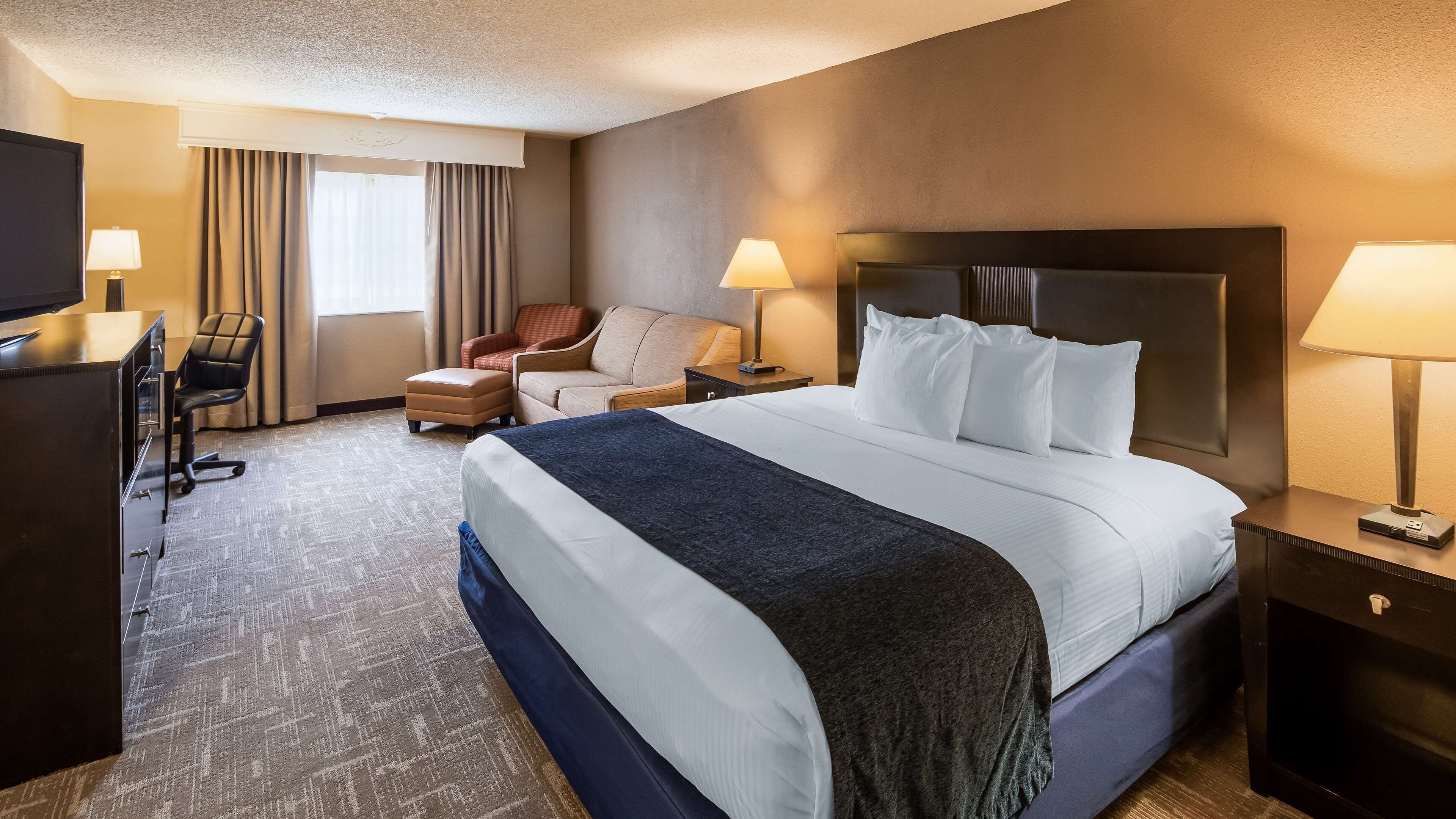 A hotel room with a large bed, two bedside tables with lamps, a desk with a chair, and a window with curtains.