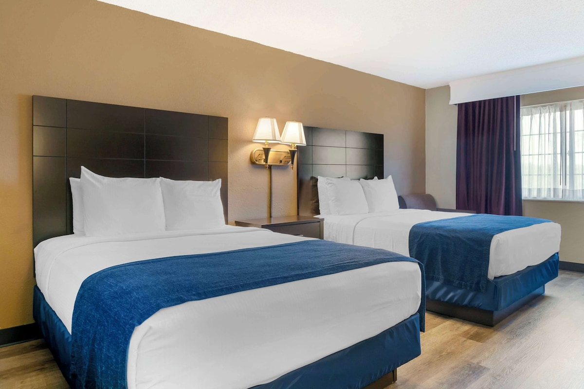 A hotel room with two beds, a nightstand with a lamp, and a window with curtains.