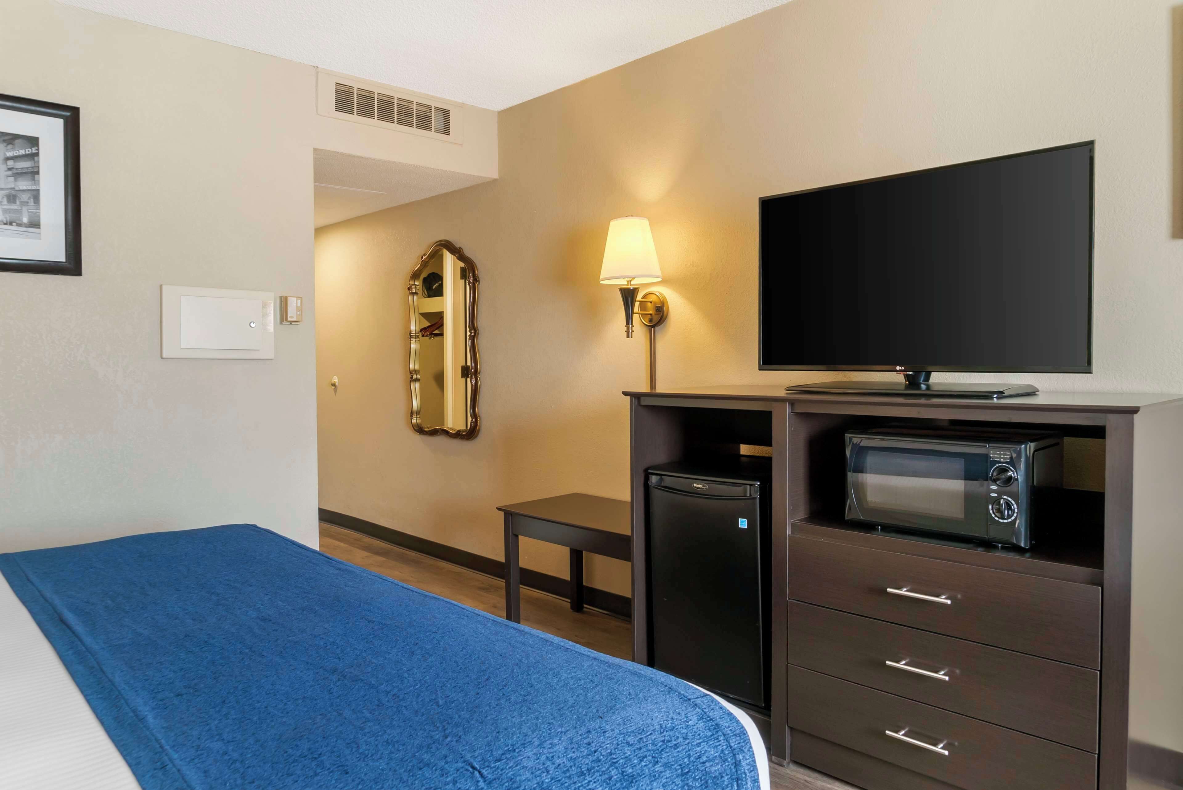 Habitación de hotel con cama, televisión, refrigerador pequeño, lámpara y espejo.