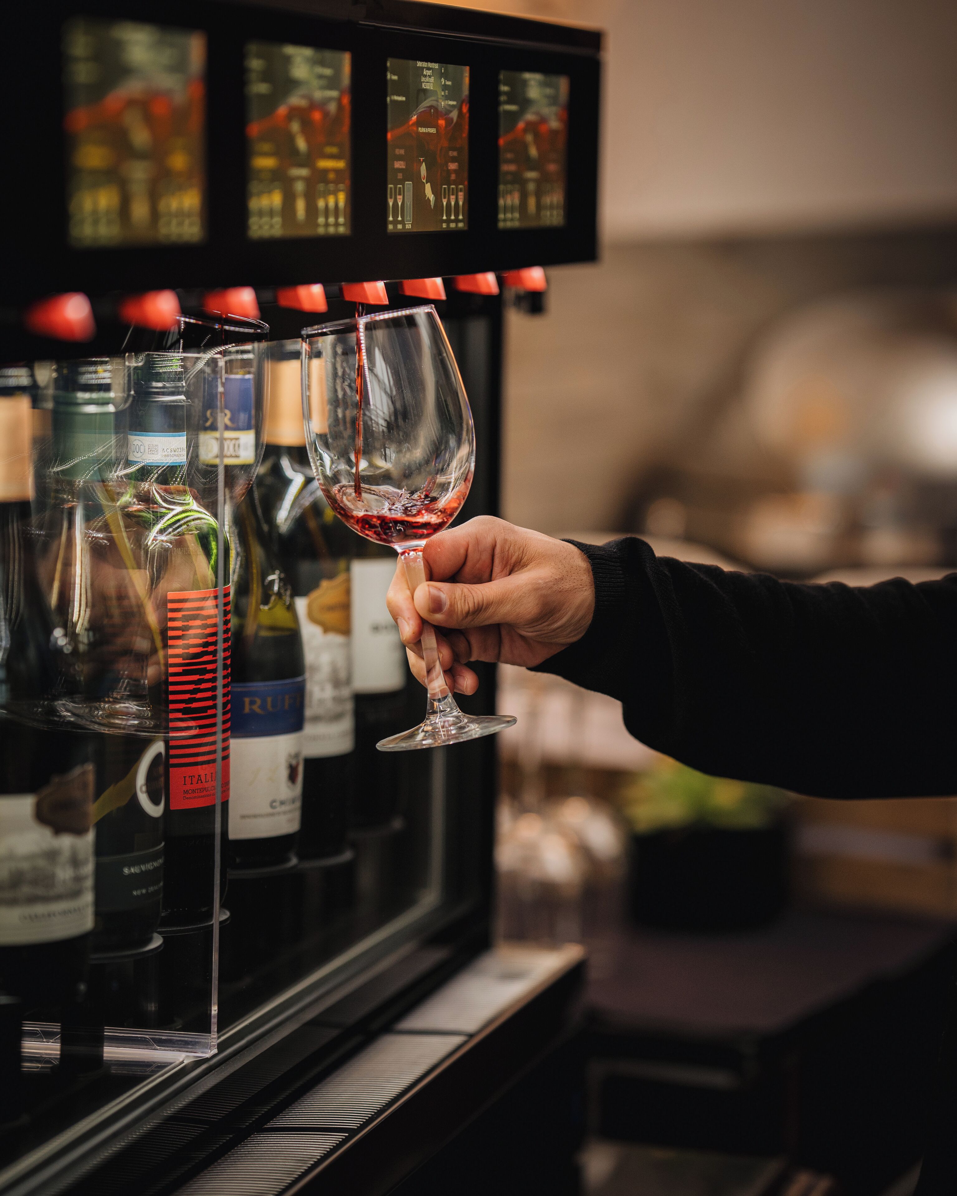 A hand holding a wine glass, with a selection of wine bottles in the background.