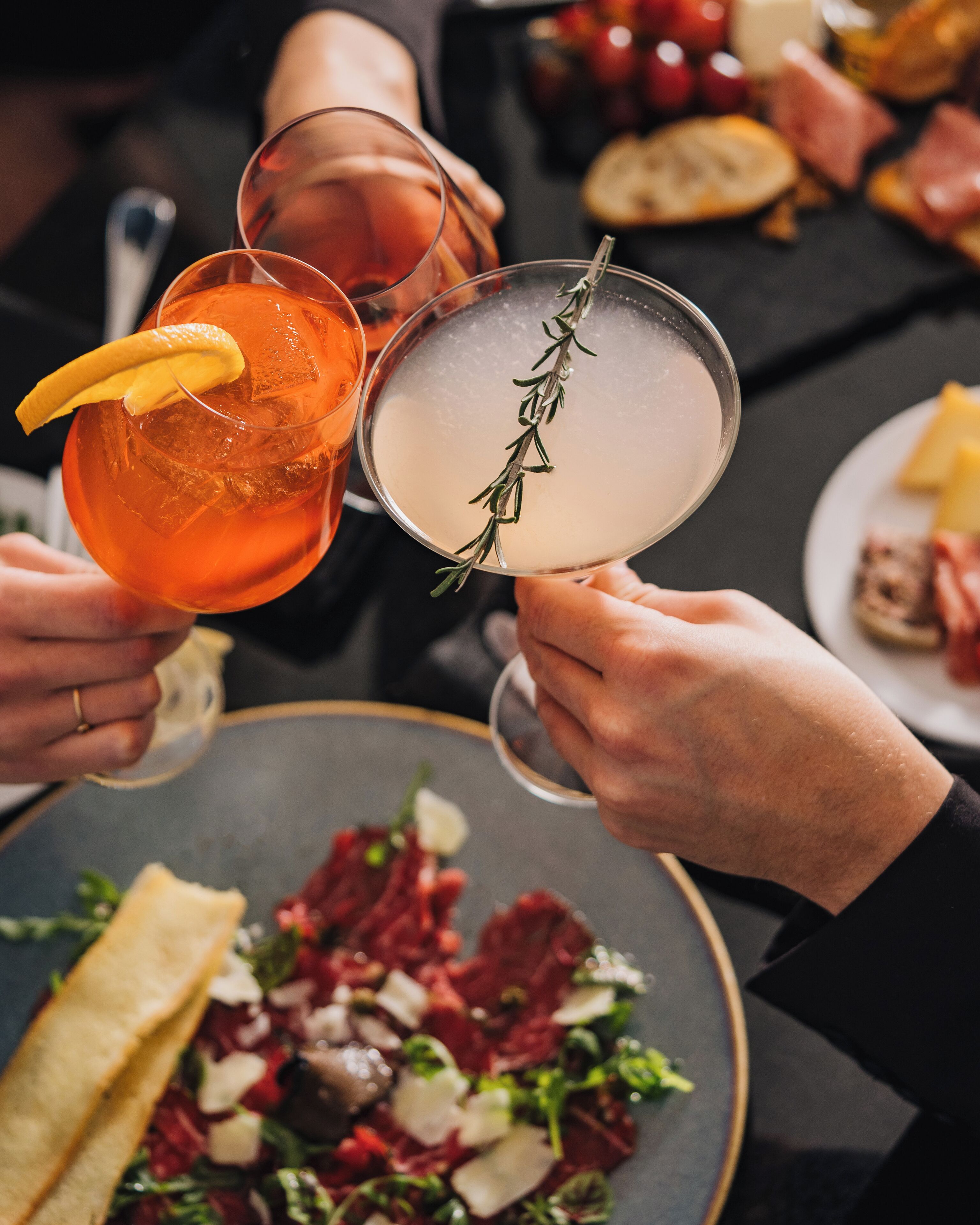 A close-up of hands holding cocktail glasses with a garnish, a plate of food, and a glass with a rosemary sprig.