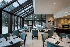 Un restaurant doté d’un plafond haut, de grandes fenêtres et d’une cheminée en pierre. Les tables sont dressées avec des assiettes, des verres et des couverts.