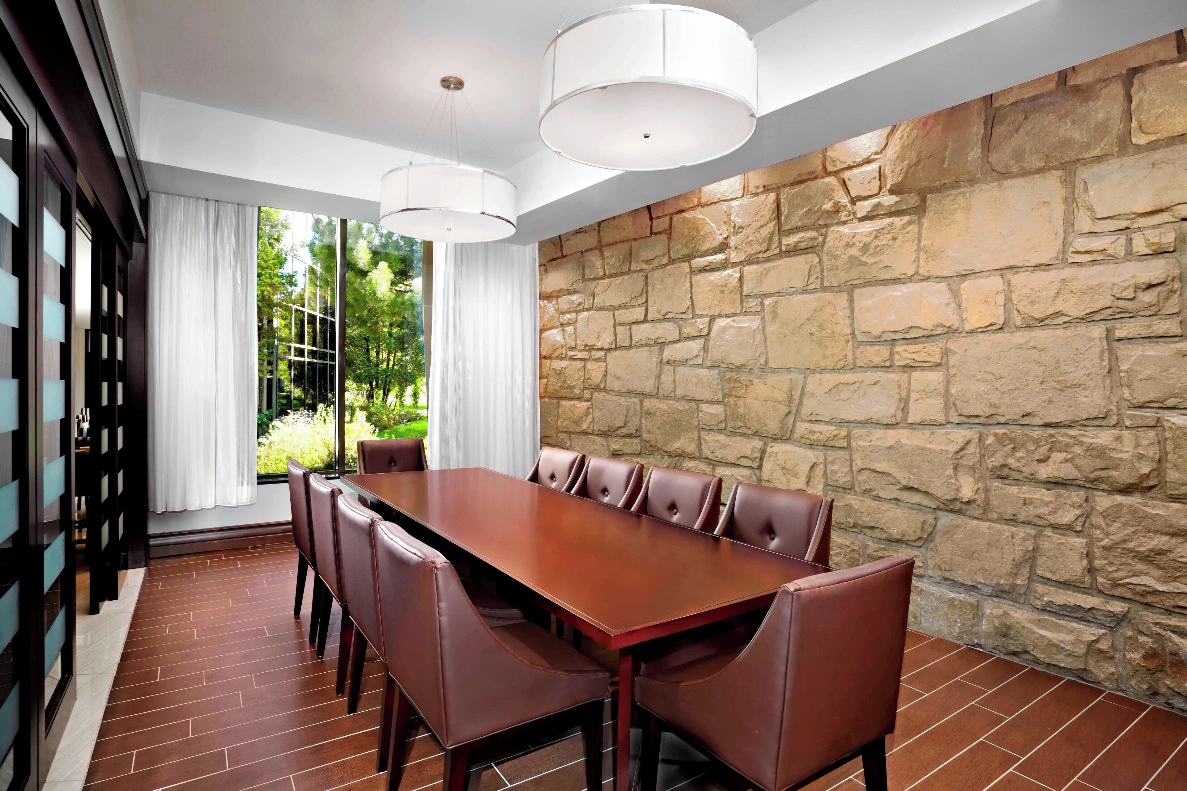 A conference room with a long table, leather chairs, a stone wall, and large windows with curtains.