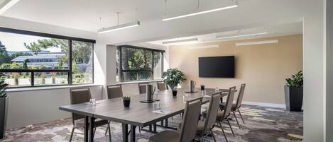 Una sala de conferencias moderna con una gran mesa, sillas, un televisor de pantalla plana y ventanas amplias con vistas a paneles solares exteriores.