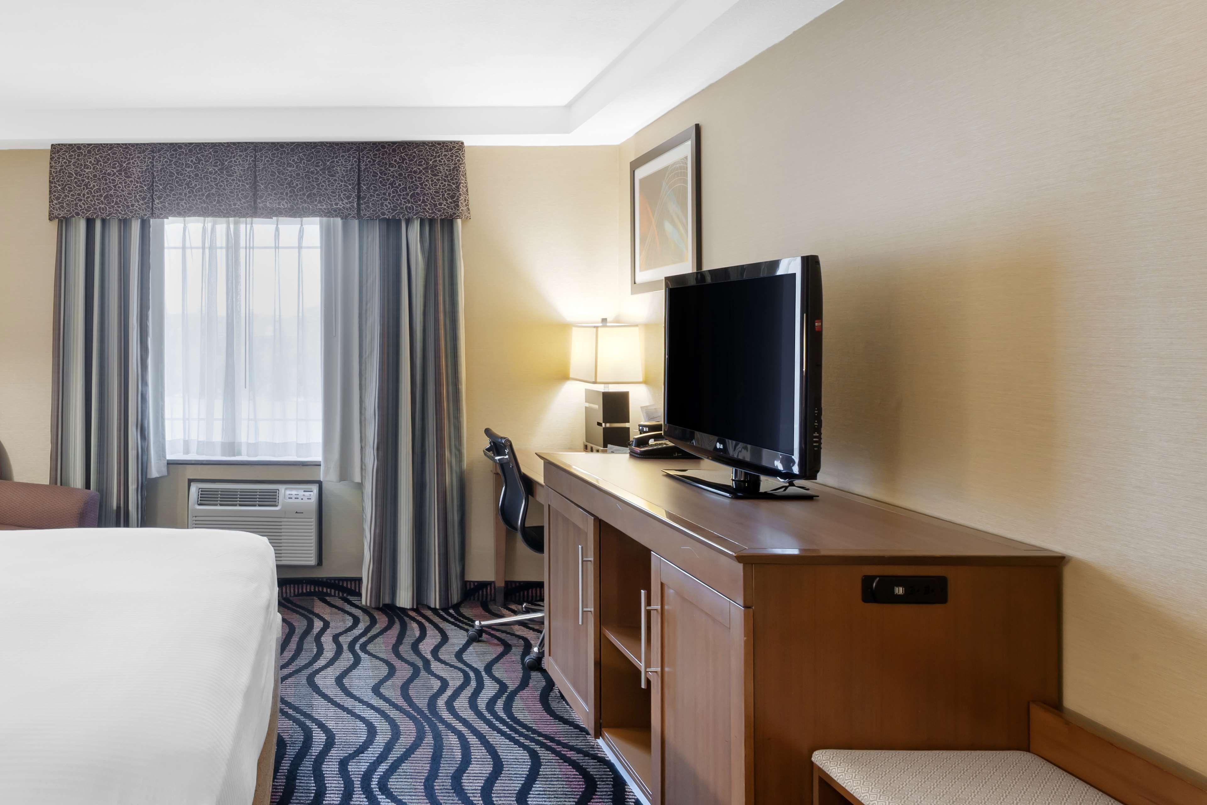 A hotel room with a bed, a television on a wooden desk, a chair, a desk lamp, and a window with curtains.