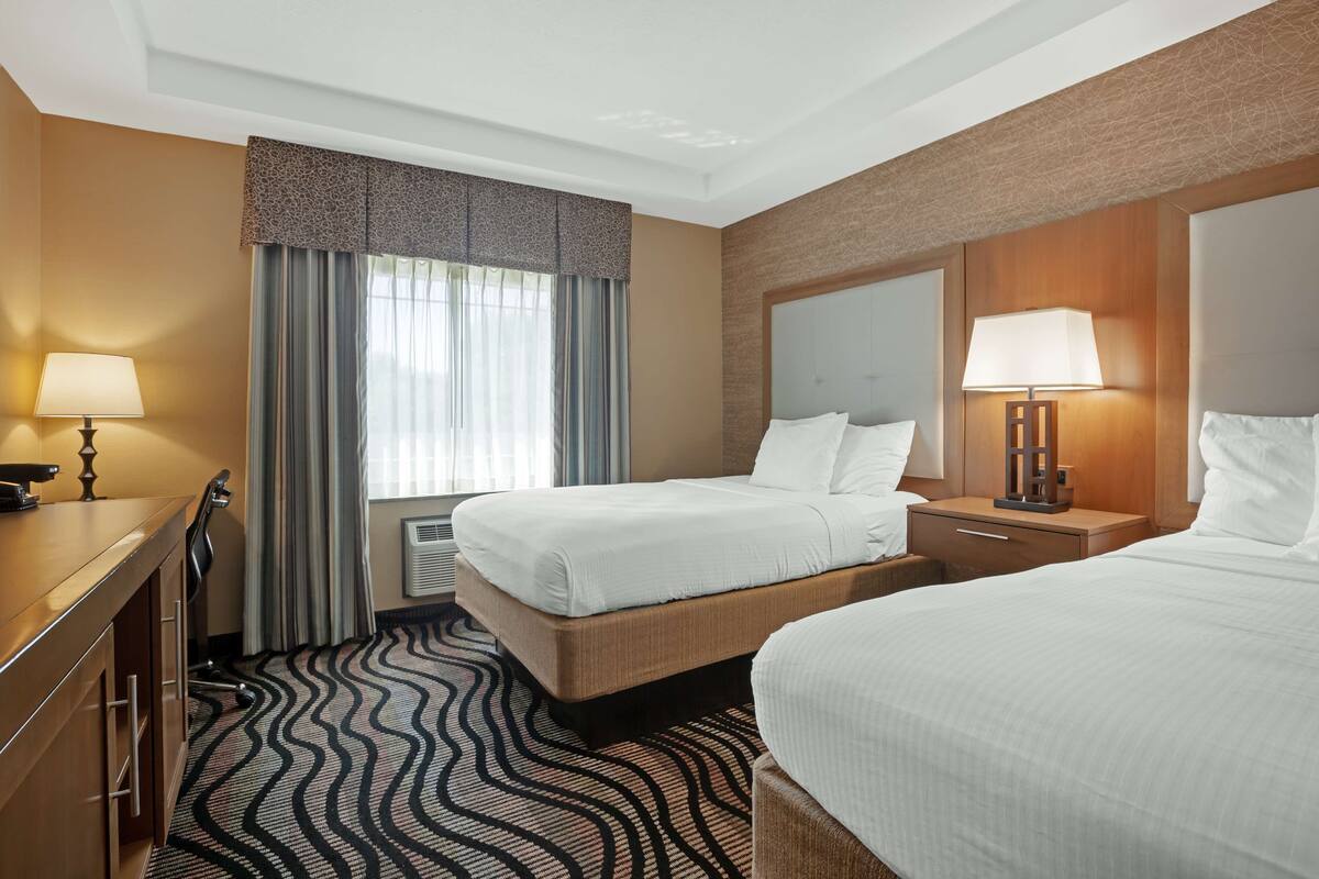 A hotel room with two beds, a desk, a lamp, a window with curtains, and a patterned carpet.