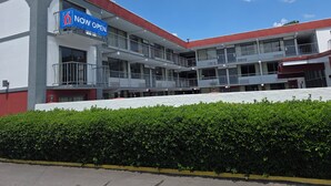 Ein mehrstöckiges Hotel mit Balkonen, einem 'NOW OPEN' Schild und einem gepflegten Heckenstreifen davor.