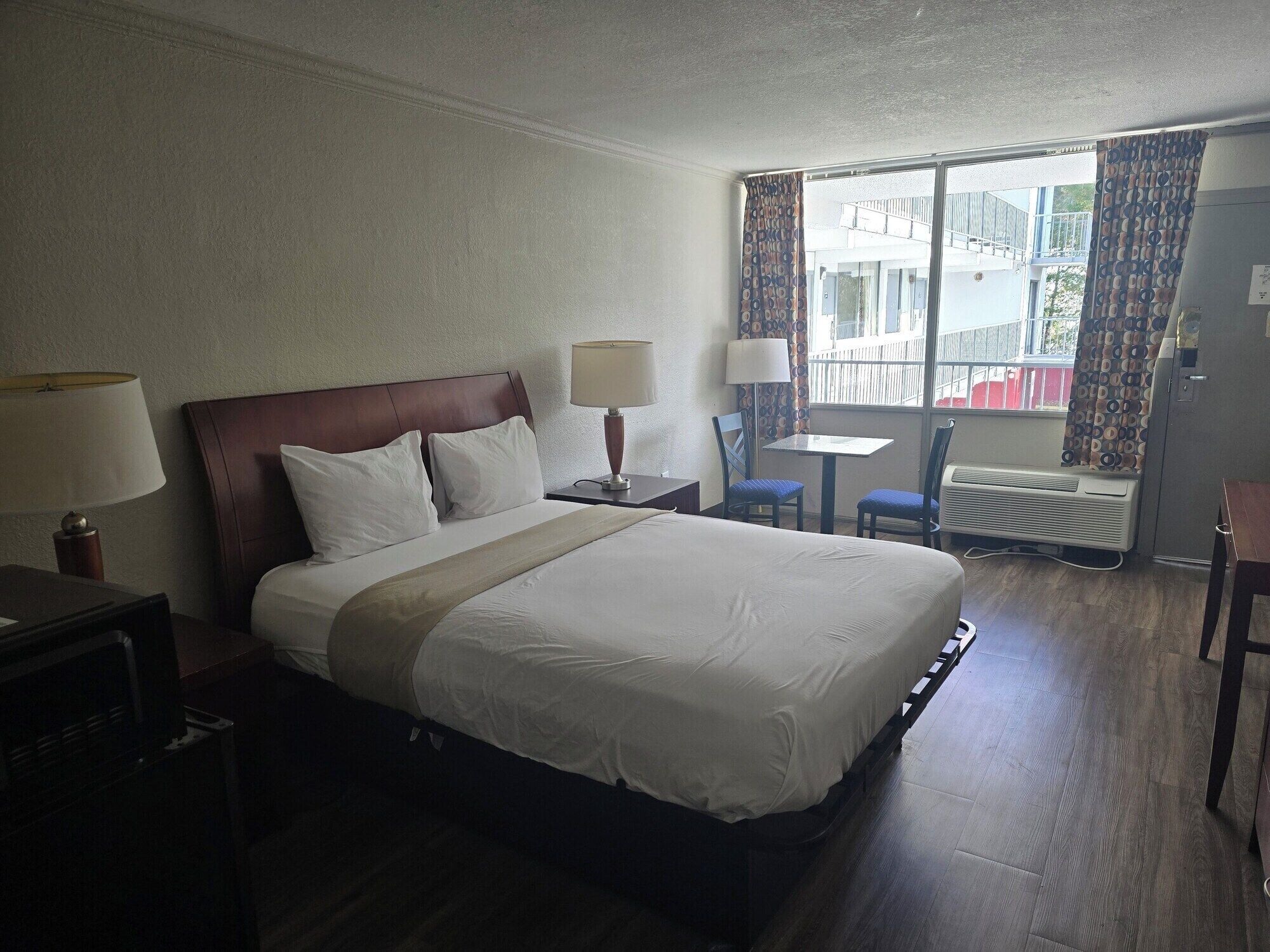 A hotel room with a large bed, two bedside lamps, a small table with chairs, a window with curtains, and a radiator.