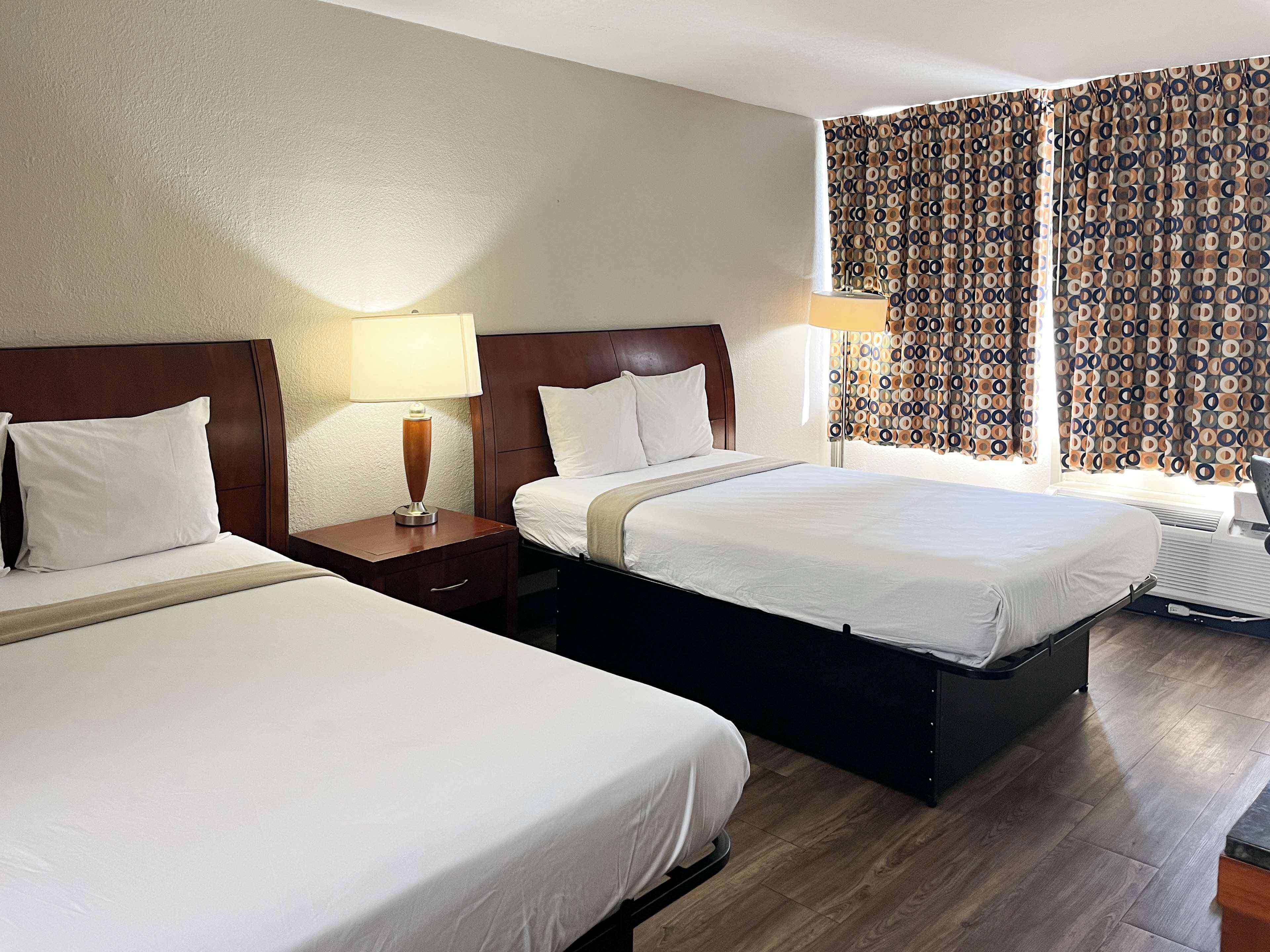 A hotel room with two beds, a nightstand with a lamp, and a window with patterned curtains.