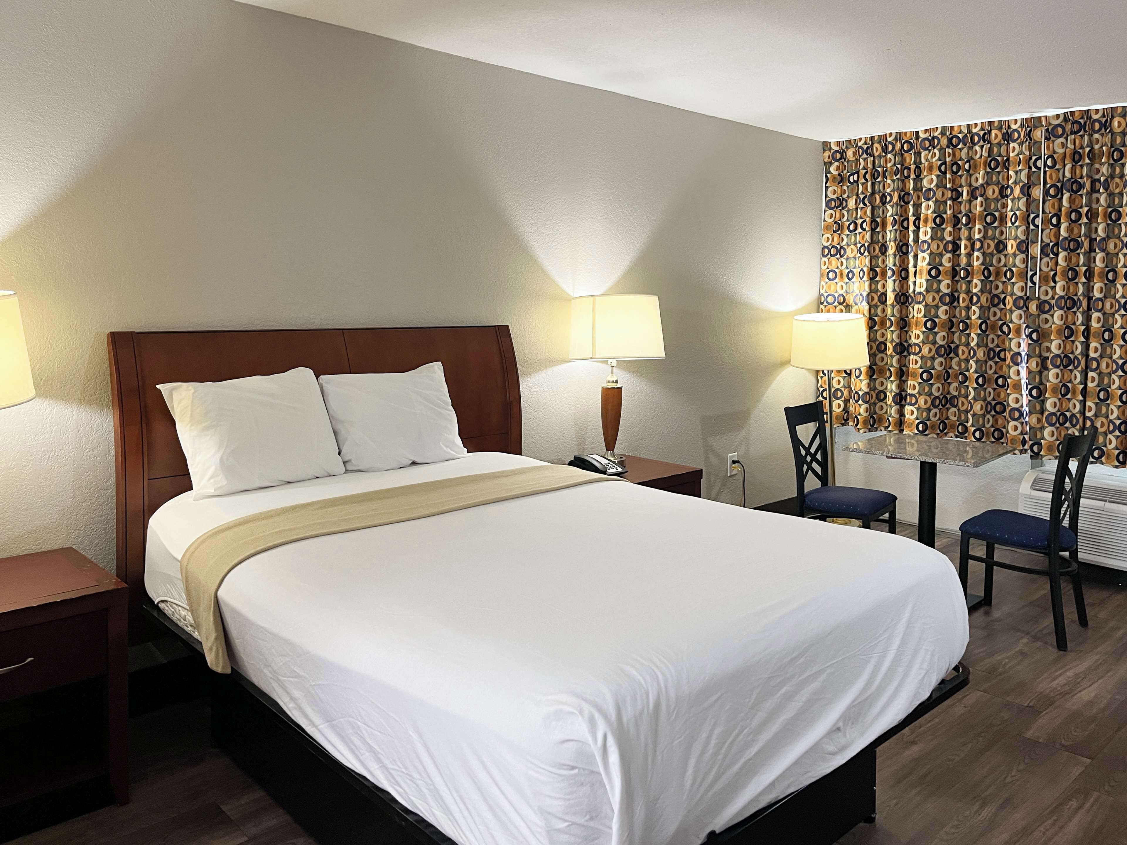 A hotel room with a large bed, two bedside tables, a desk with a chair, patterned curtains, and a window.