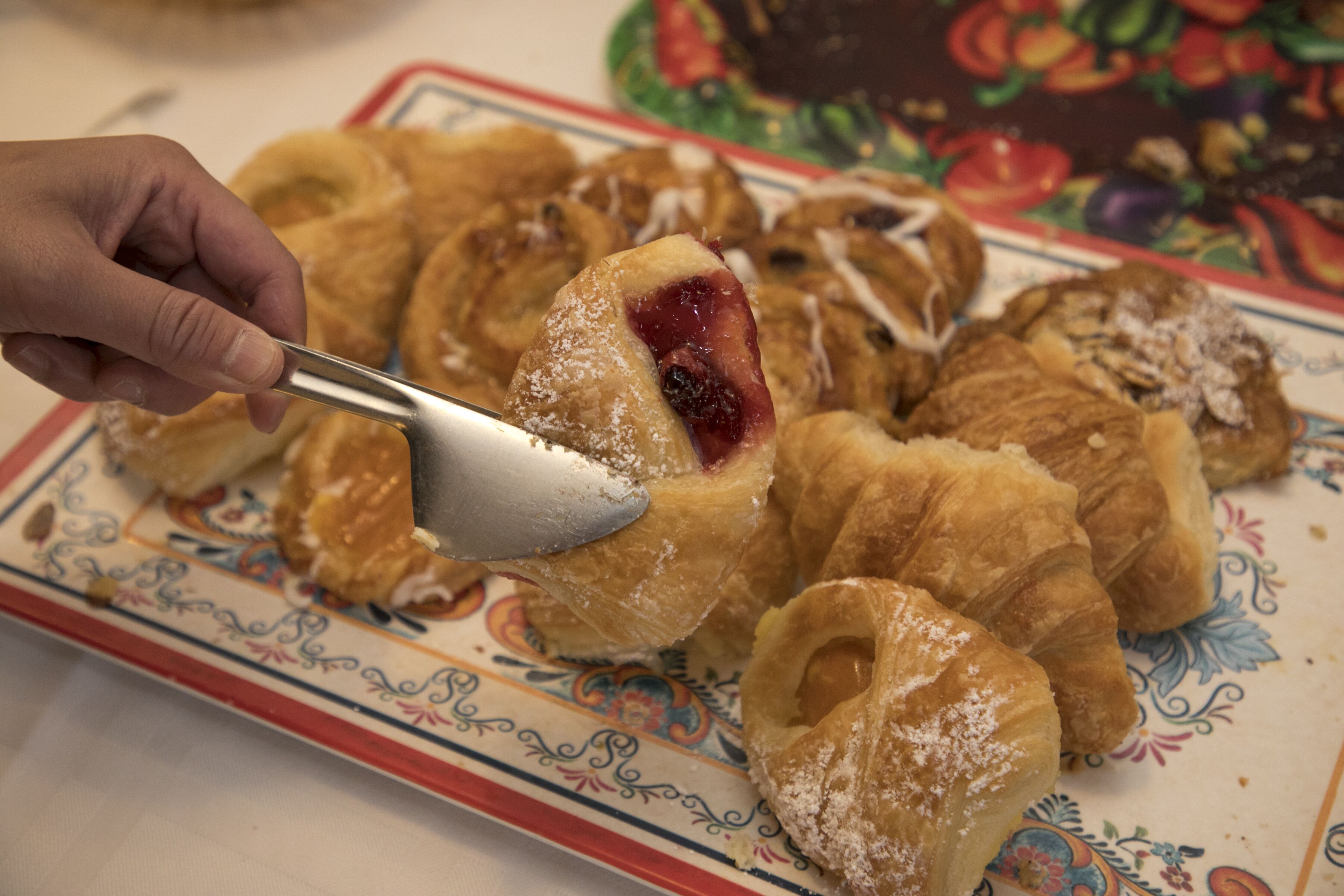 Een hand die met een lepel boven een bord gebak staat, waaronder croissants en een gebakje met rode vulling.