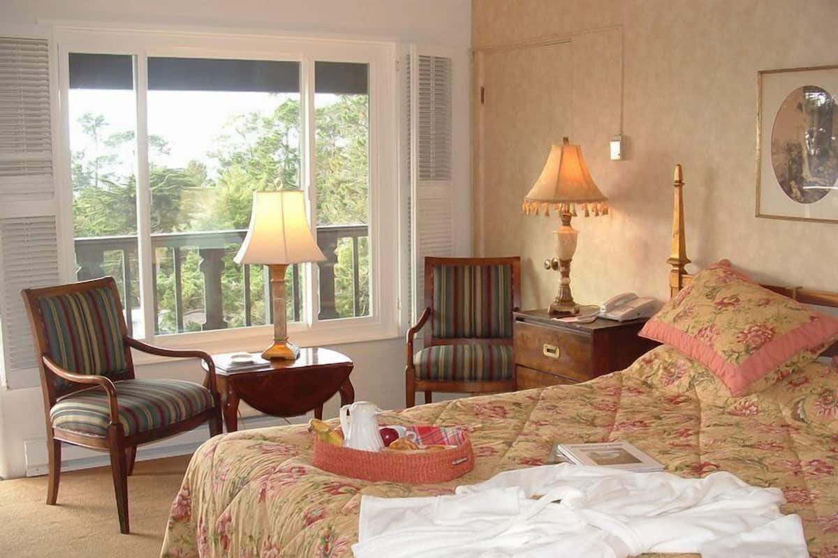 A bedroom with a large bed, two chairs, a nightstand, a lamp, and a window with a view of trees.