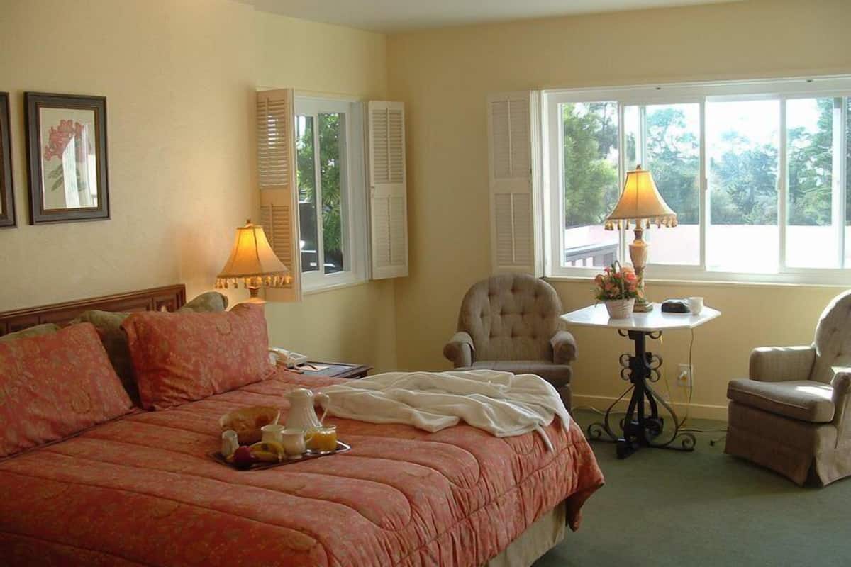 A hotel room with a large bed, two chairs, a small table with a lamp, and a window with shutters.