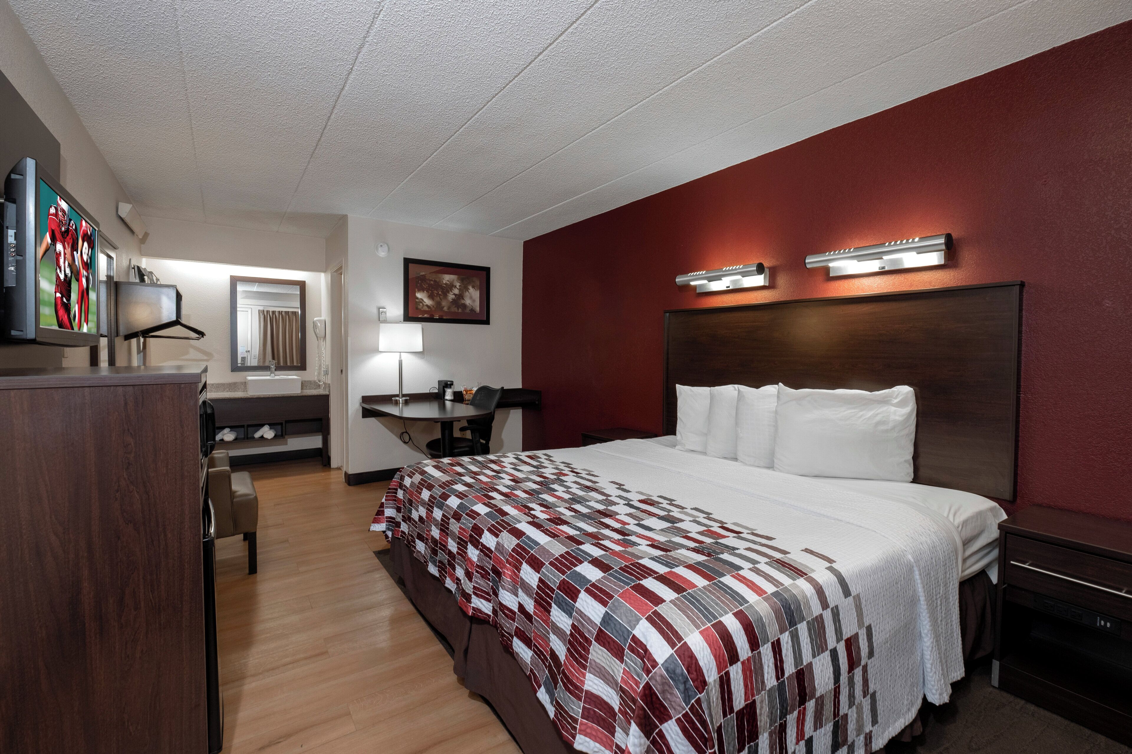 A hotel room with a large bed, a desk with a chair, a television, a mirror, and a red accent wall.