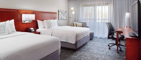 A hotel room with two beds, a desk, a chair, a TV, and a large window with curtains.
