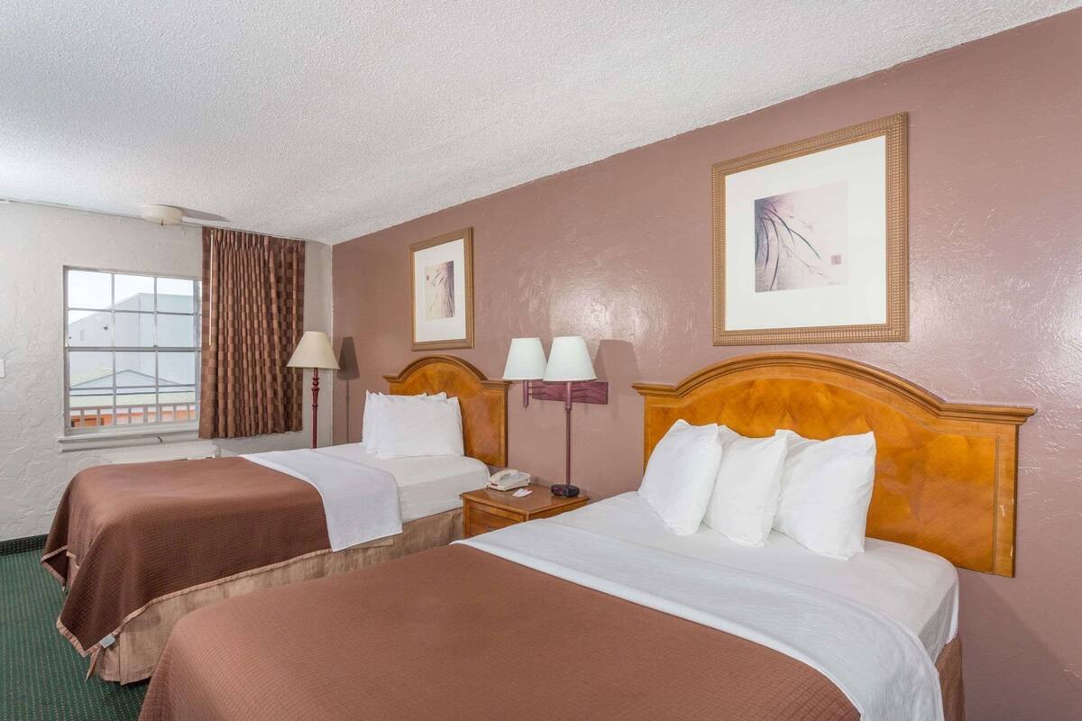 Een hotelkamer met twee bedden, een raam met gordijnen, een nachtkastje met een lamp en twee ingelijste foto’s aan de muur.