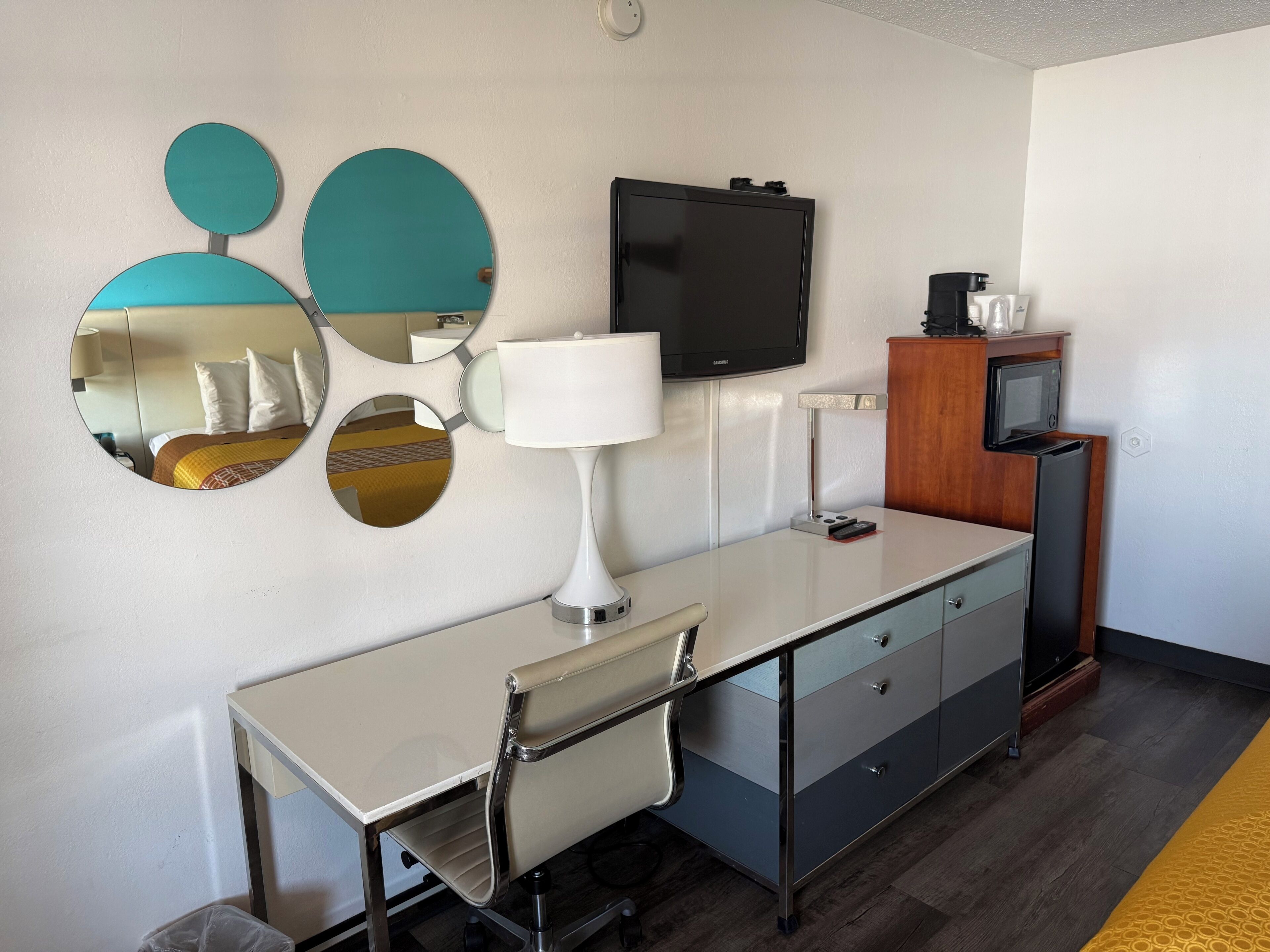 A hotel room with a desk, chair, TV, mirrors, and a minibar.