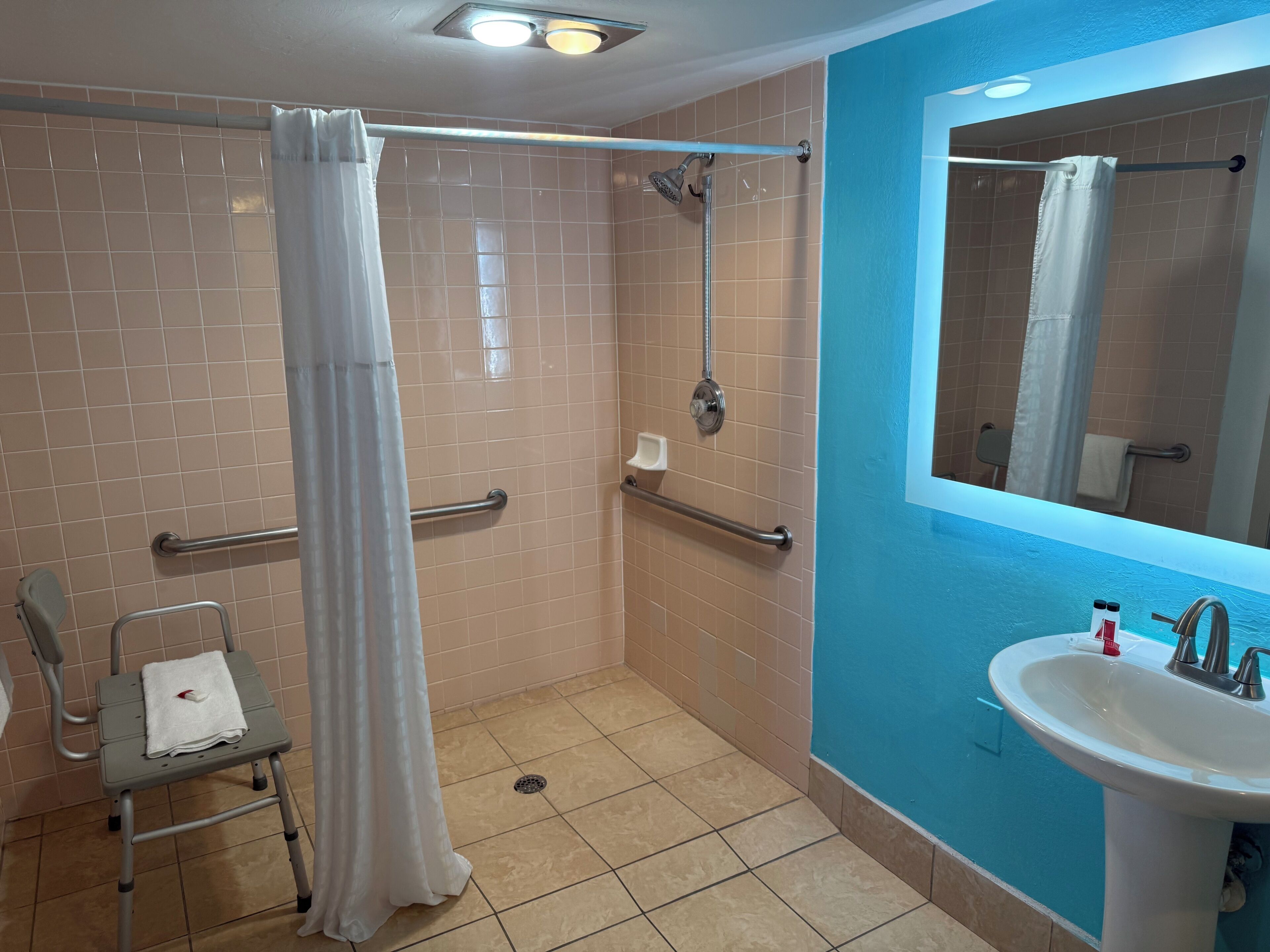 A bathroom with a shower, a handrail, a sink, and a wheelchair-accessible chair.