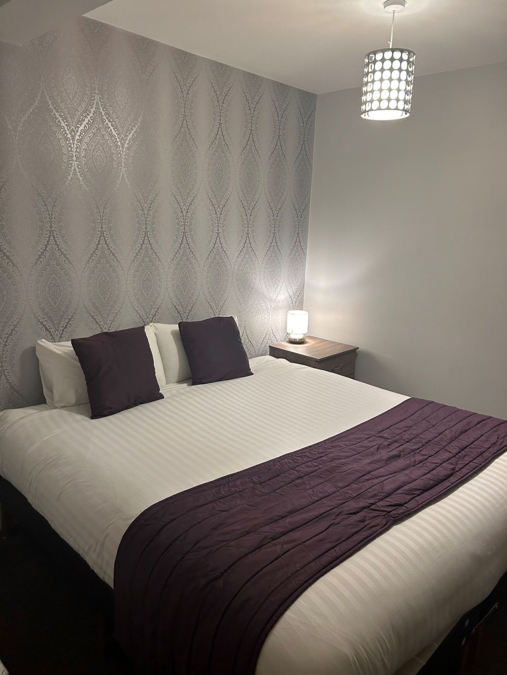 Un lit bien fait, avec une couvre-lit blanche et violette, une tête de lit sombre et un papier peint à motifs.