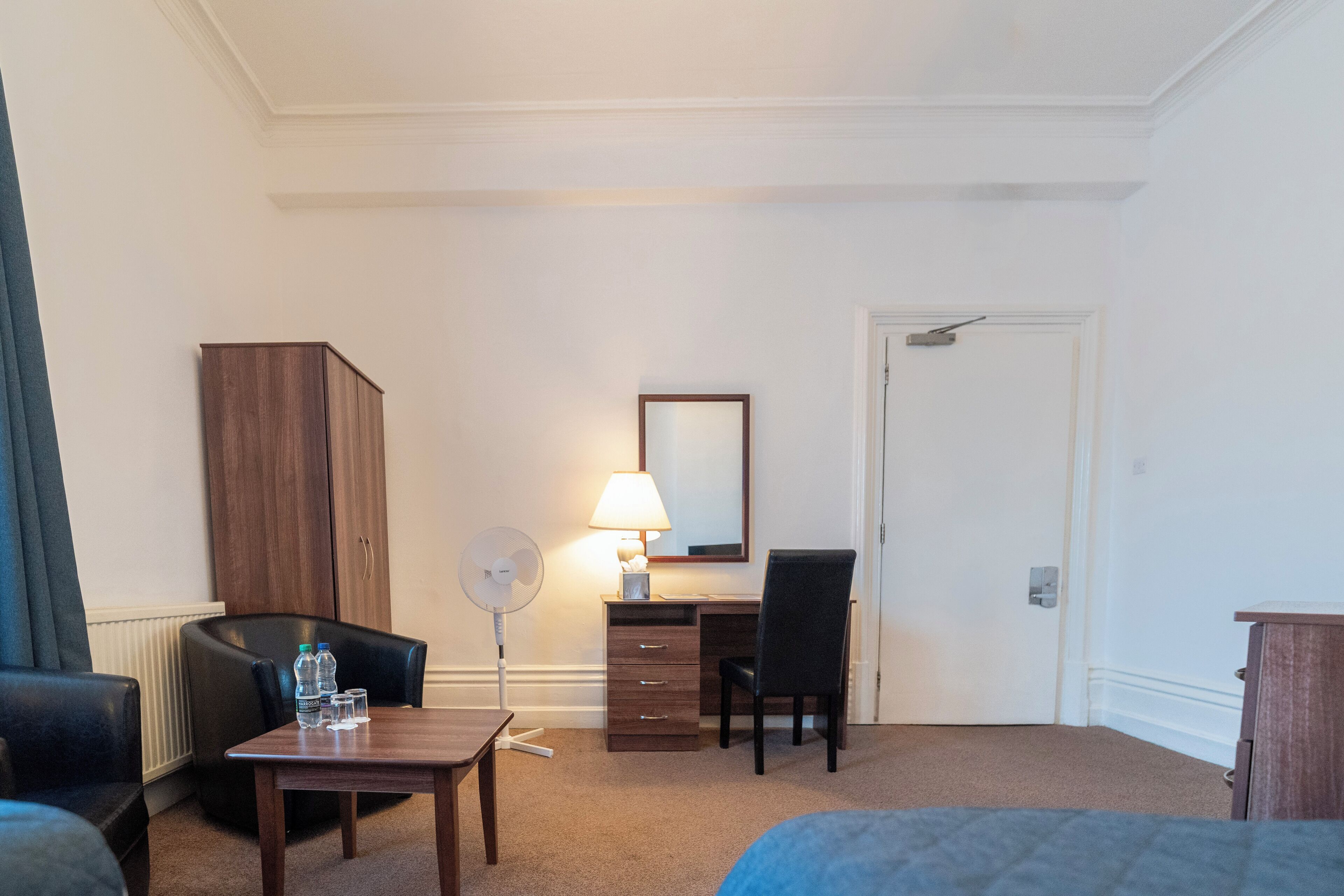 A hotel room with a bed, a desk, a chair, a wardrobe, a lamp, and a fan.