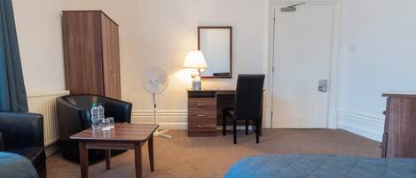 A hotel room with a bed, a desk, a chair, a wardrobe, a lamp, and a fan.