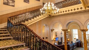 Una imponente escalera con barandales ornamentales y un candelabro, que conduce a un balcón en el segundo piso con balaustrada.