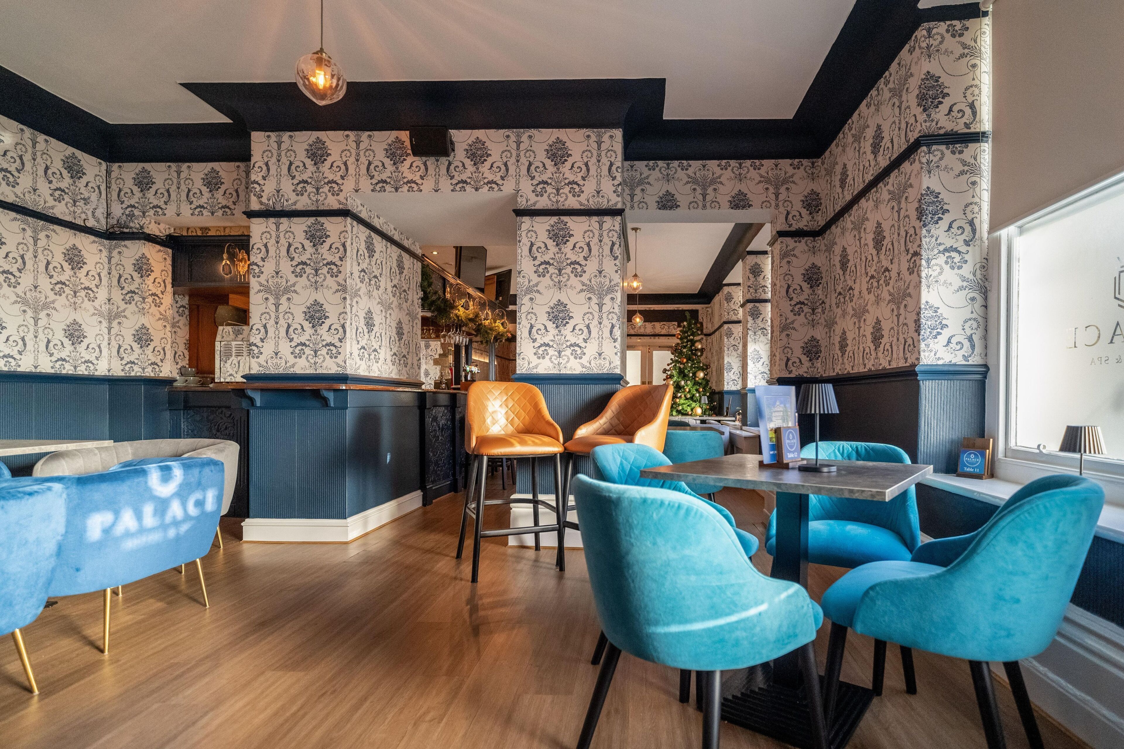 A restaurant interior with blue and white patterned wallpaper, wooden flooring, and blue and orange chairs.