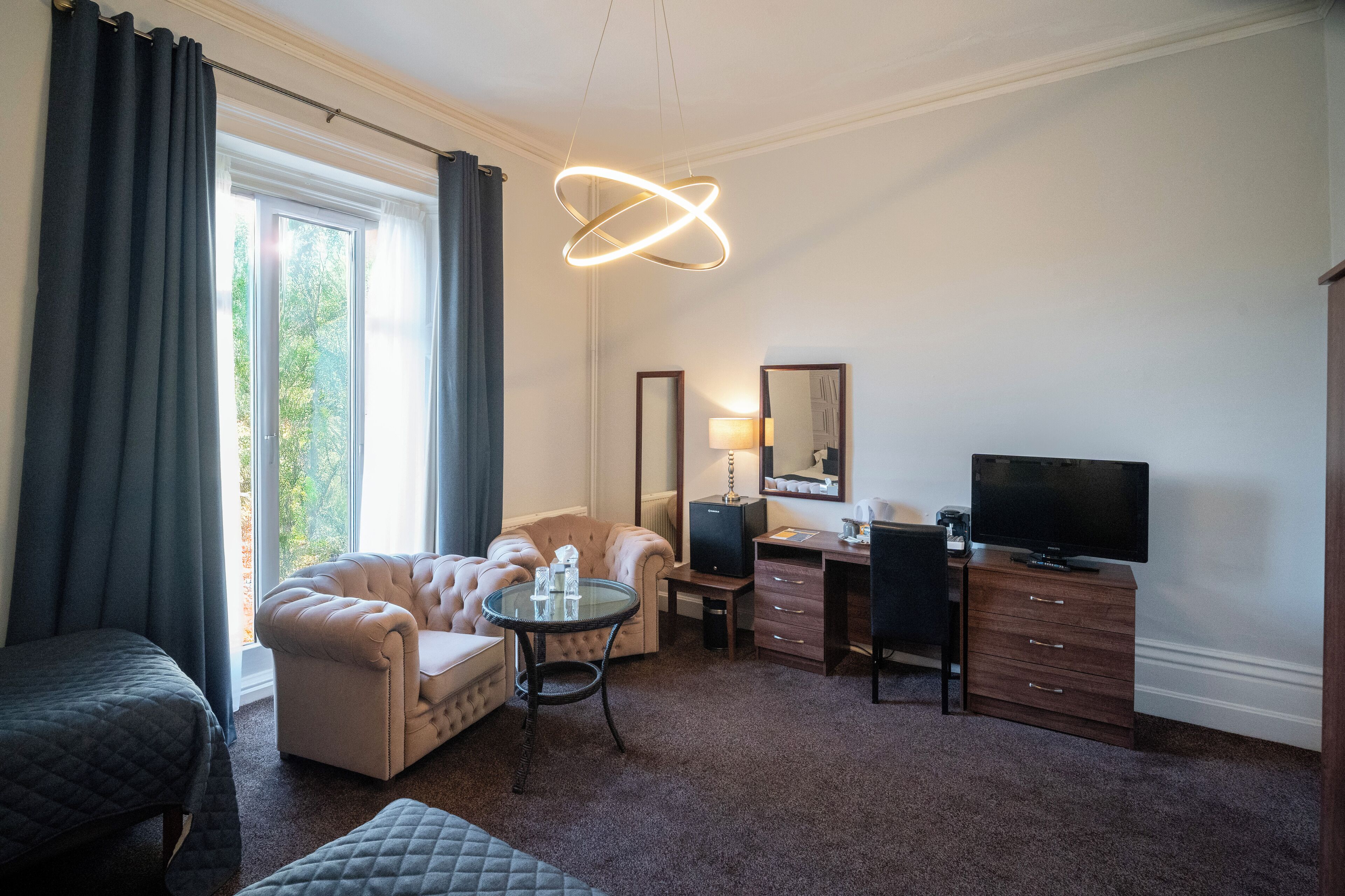 A hotel room with a bed, a seating area with a sofa and armchair, a desk with a computer and TV, and a large window with curtains.