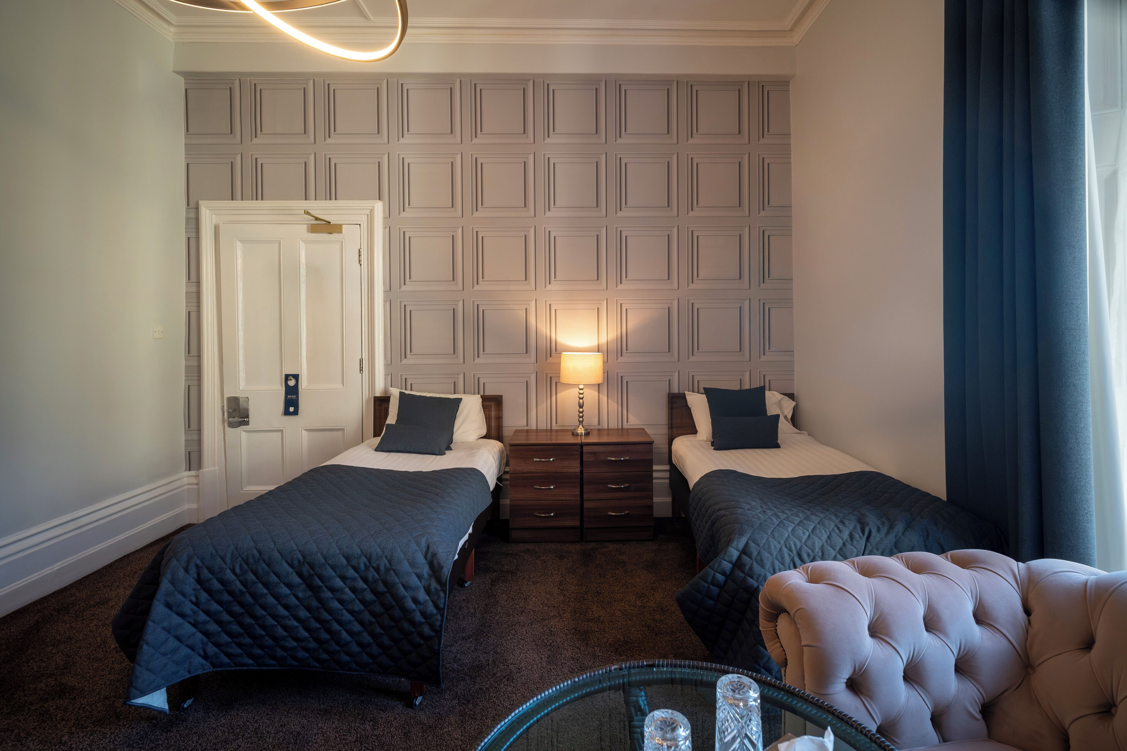 A hotel room with two beds, a nightstand, a lamp, a chair, and a door.